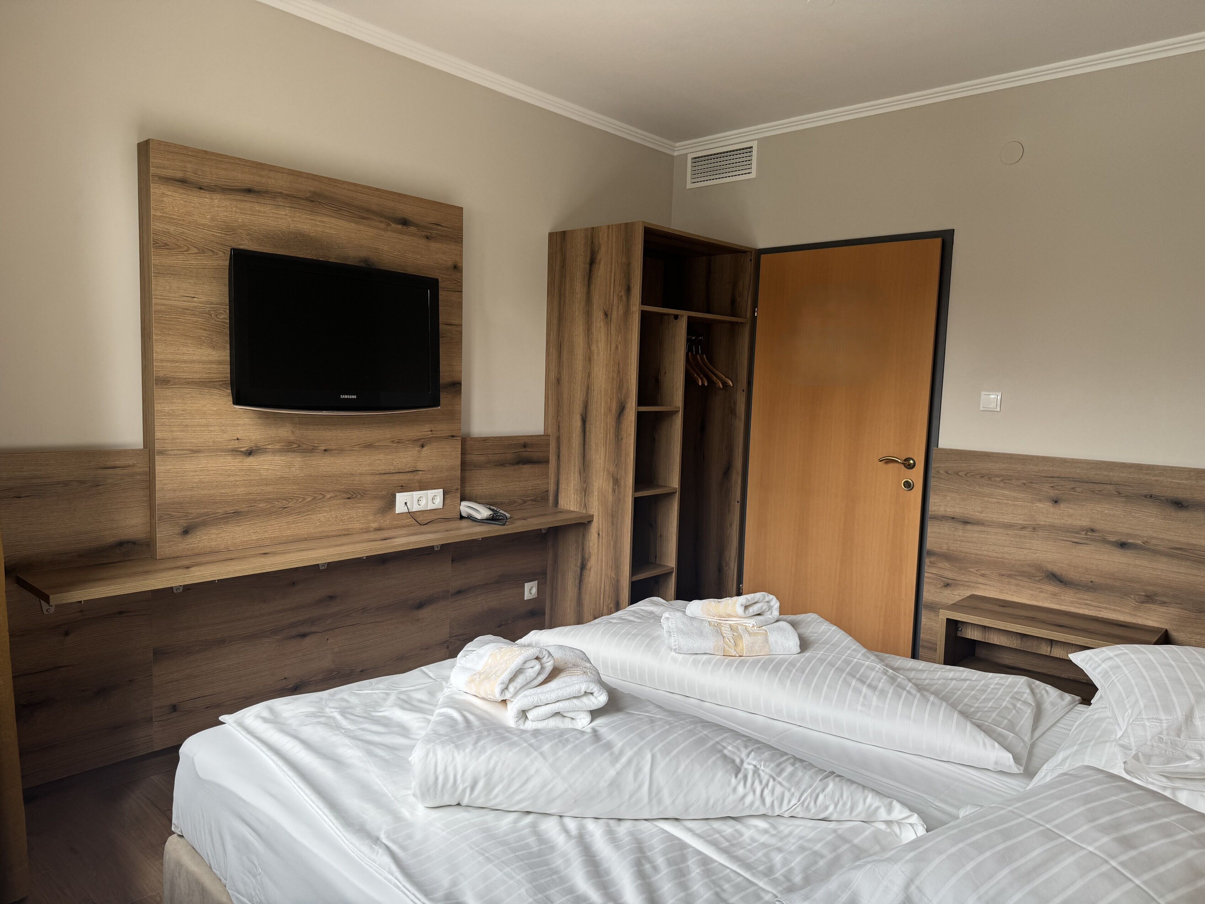 A bedroom with a wooden headboard, a flat-screen TV, a wardrobe, and a bed with neatly made white linens and towels.