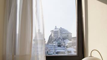 A room with a desk, a lamp, a laptop, and a view of a building through a window with sheer curtains.