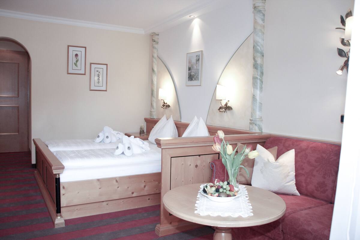 A hotel room with a bed, a red sofa, a round table with a white tablecloth, and wall-mounted lamps.