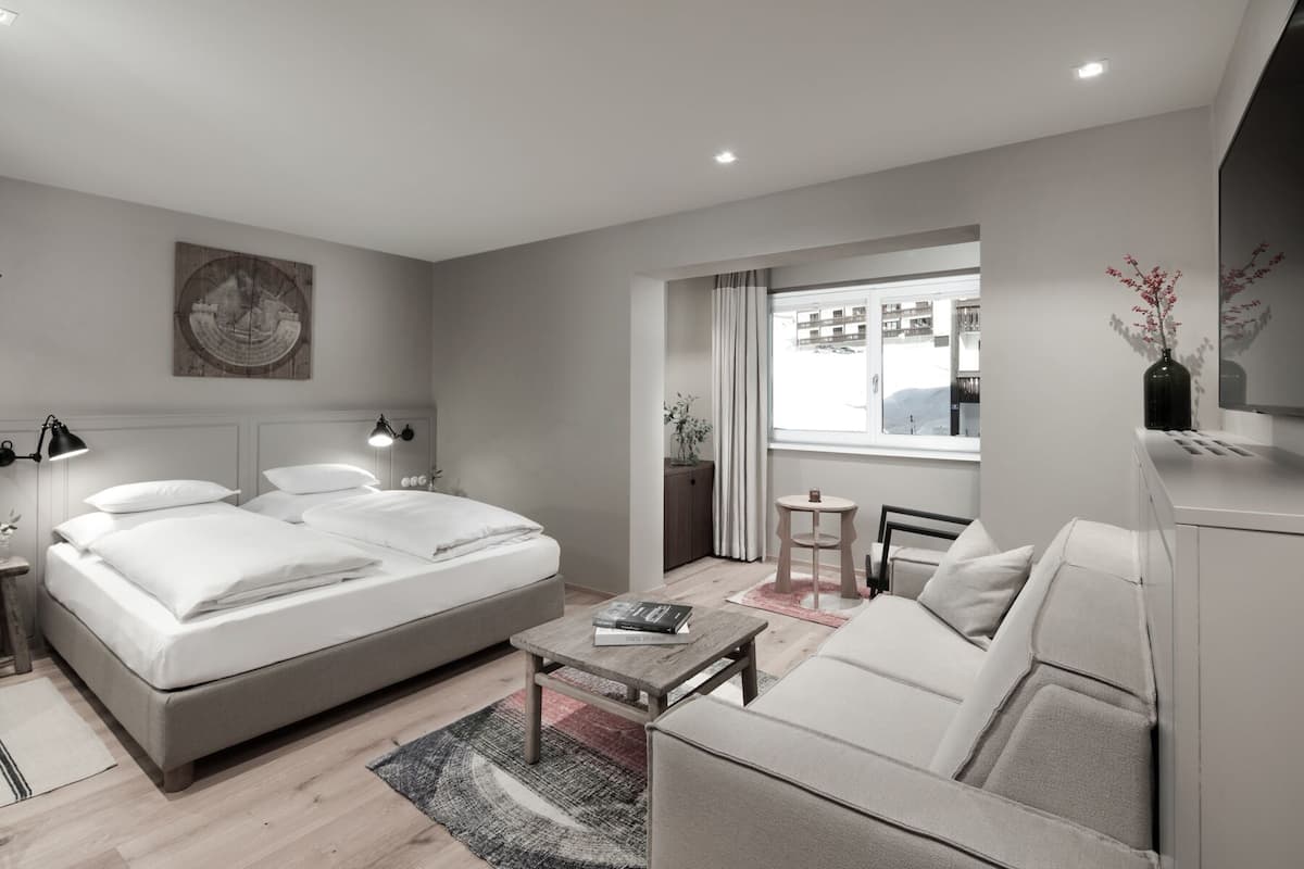 A modern hotel room with a large bed, a sofa, a small table, and a view of the city through the window.