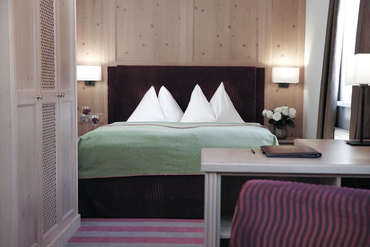 A hotel room with a bed, bedside lamps, a small table with a notebook, a vase with flowers, and a striped chair.