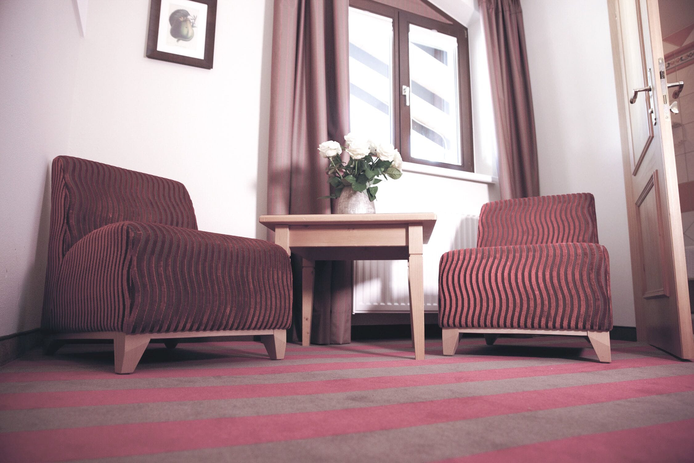 A room with two striped armchairs, a small wooden table with a vase of flowers, and a window with curtains.