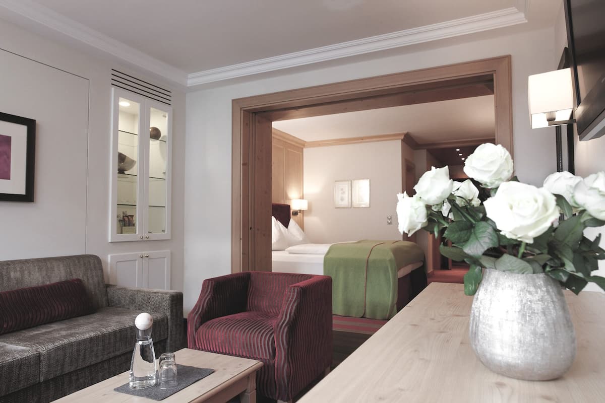 A hotel room with a sofa, armchair, and a table with a vase of flowers. There is a bed with a green blanket and a bedside table with a lamp.