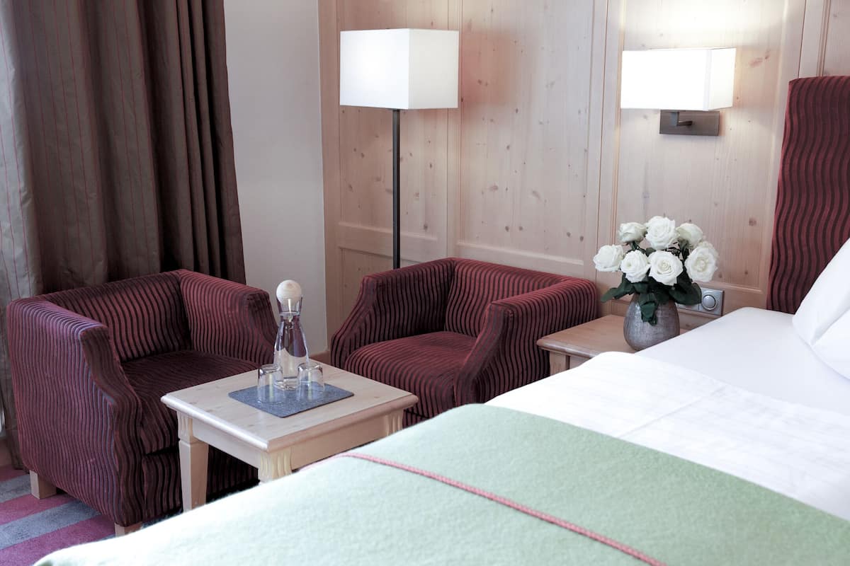 A hotel room with a bed, two armchairs, a small table with a vase of flowers, and two wall-mounted lamps.