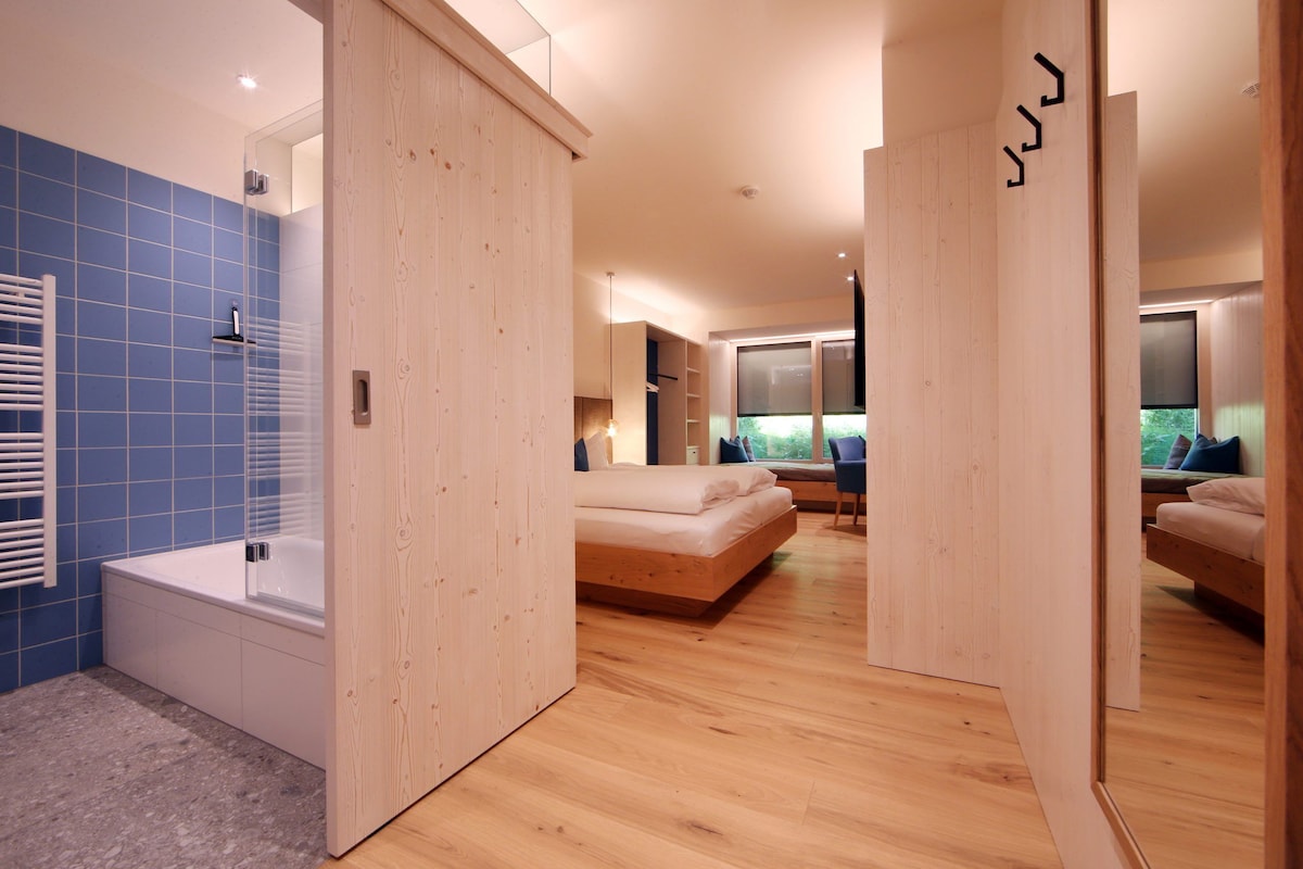 Un dormitorio moderno con suelo de madera, una cama grande, una zona de descanso separada y un baño con azulejos azules.