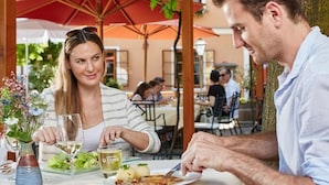 Una pareja cenando en un café al aire libre con mesas, sombrillas y una vista de un edificio al fondo.