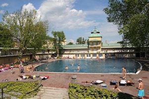 Zona de piscina con un edificio grande al fondo, rodeada de árboles y gente disfrutando de las instalaciones.