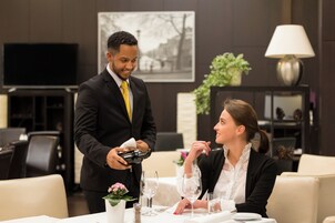Un hombre con traje sirve una bebida en un vaso, sentado a una mesa frente a una mujer. Se ve una televisión al fondo, una planta y una lámpara en una mesita auxiliar.