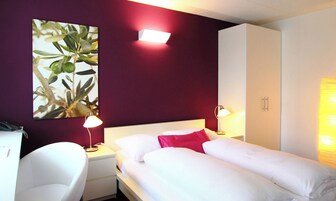 Una habitación de hotel con una cama, dos lámparas de noche, una mesita de noche, un escritorio con una silla, un armario y una obra de arte grande en la pared.