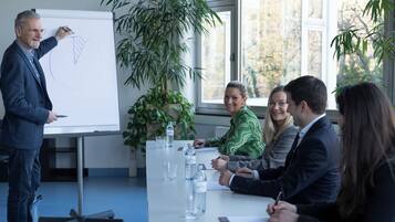 A business meeting with a presenter and four attendees around a table with a whiteboard, in a room with large windows and indoor plants.