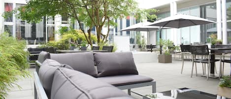 An outdoor seating area with a grey sofa, a glass-top table, and umbrellas.