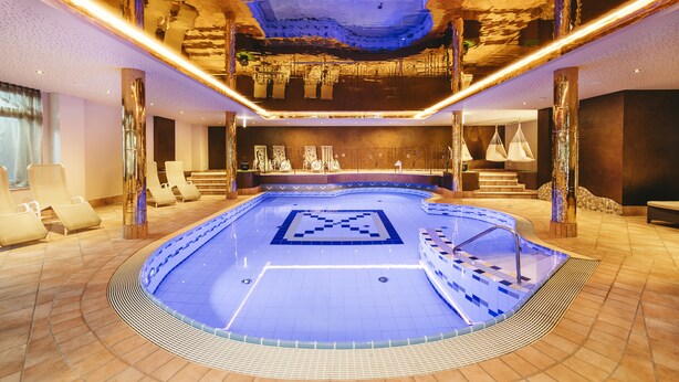 Piscina interna com padrão geométrico de azulejos, cercada por cadeiras de descanso e um design de teto moderno.