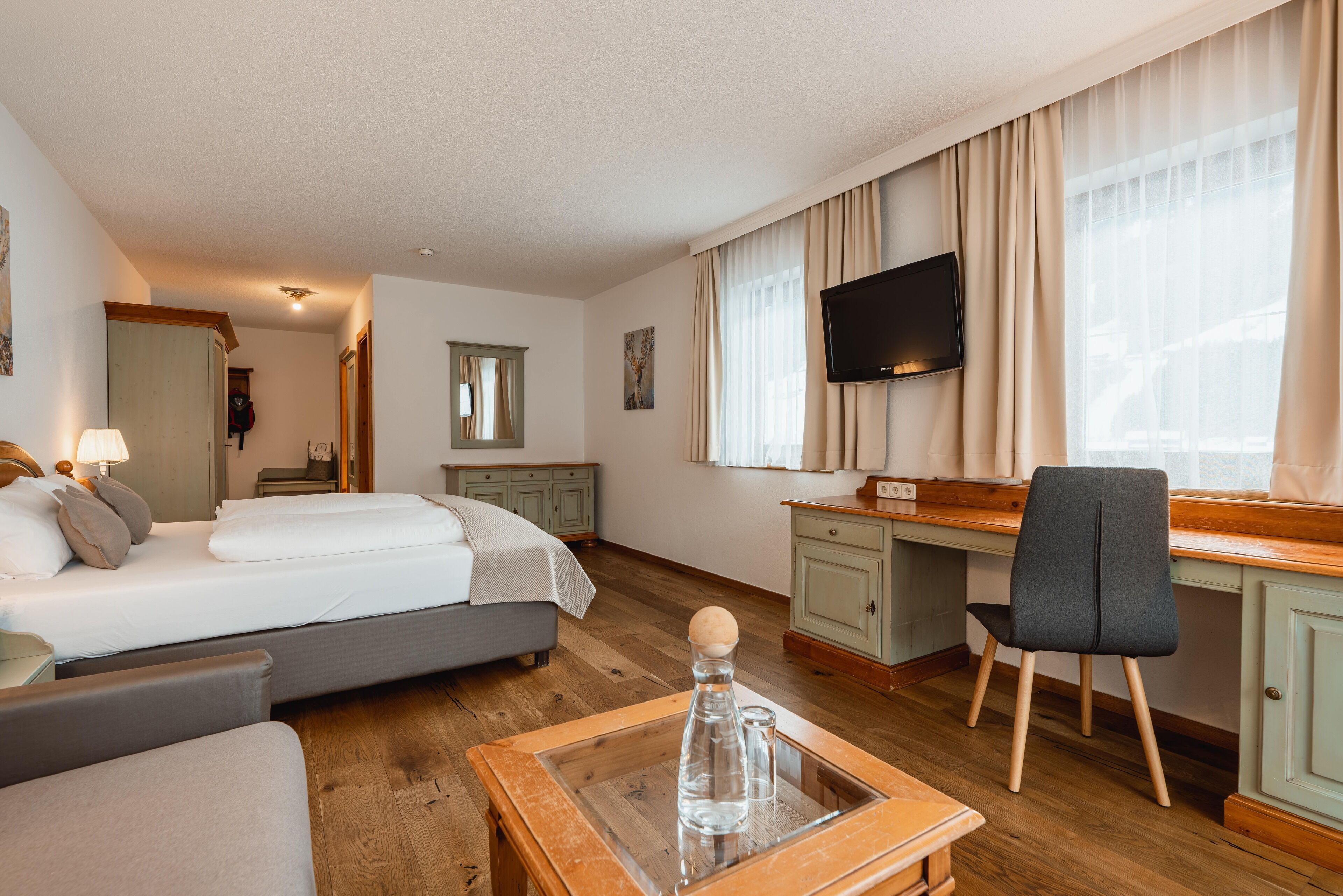 A hotel room with a bed, desk, chair, TV, and a small table with a bottle and glasses.