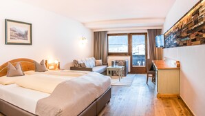 Quarto de hotel com uma cama, um sofá, uma mesinha, uma cadeira, uma televisão e uma prateleira na parede.
