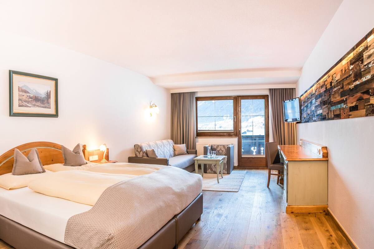 A hotel room with a bed, a sofa, a small table, a chair, a TV, and a wall-mounted shelf.