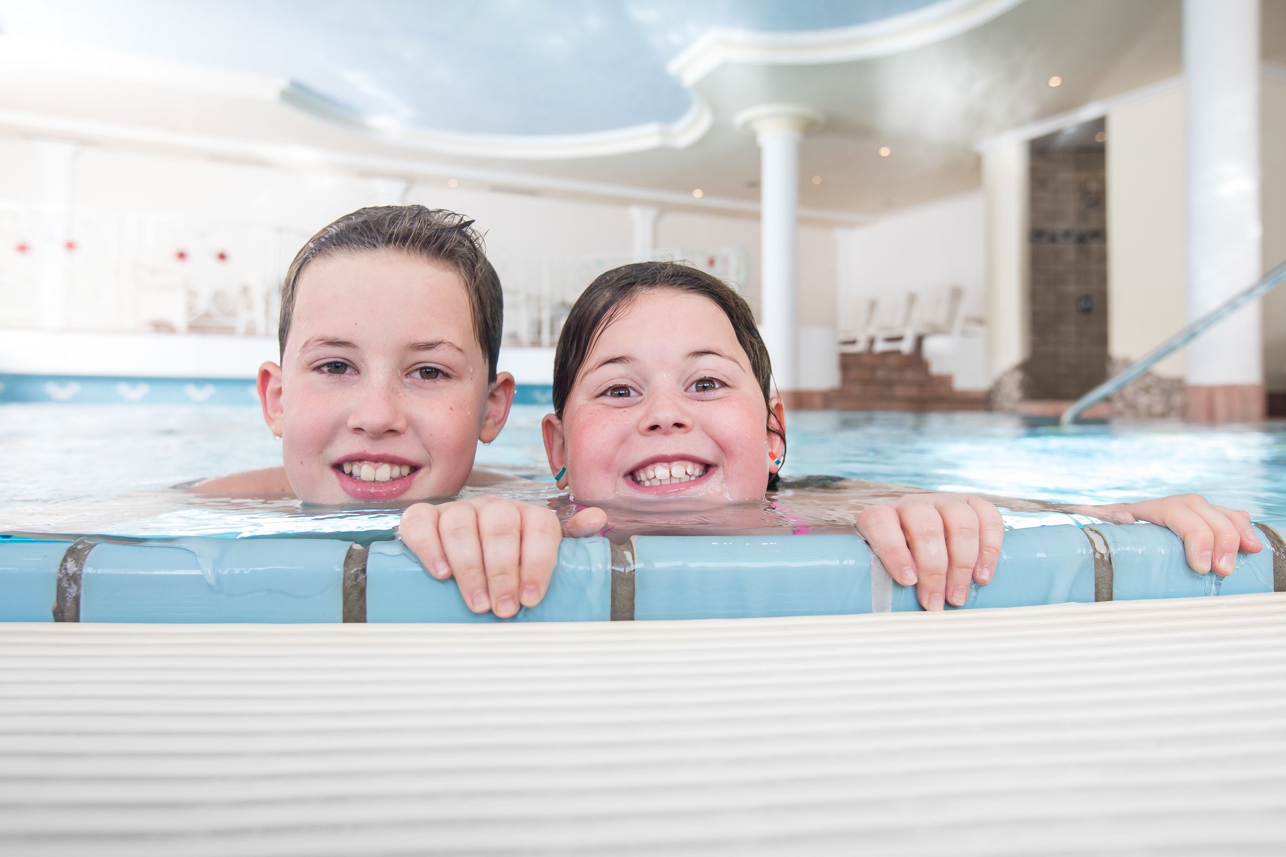 Two children in a pool with a modern indoor setting, including a staircase and a clear ceiling.