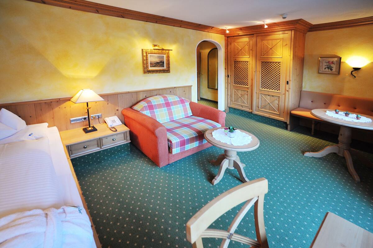 A hotel room with a bed, a sofa, a small table, and a chair. There is a lamp on the table and a picture on the wall.