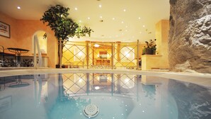 An indoor swimming pool with a clear water surface, surrounded by a lounge area with seating and a large tree. - Sporthotel Kirchler (Tux)