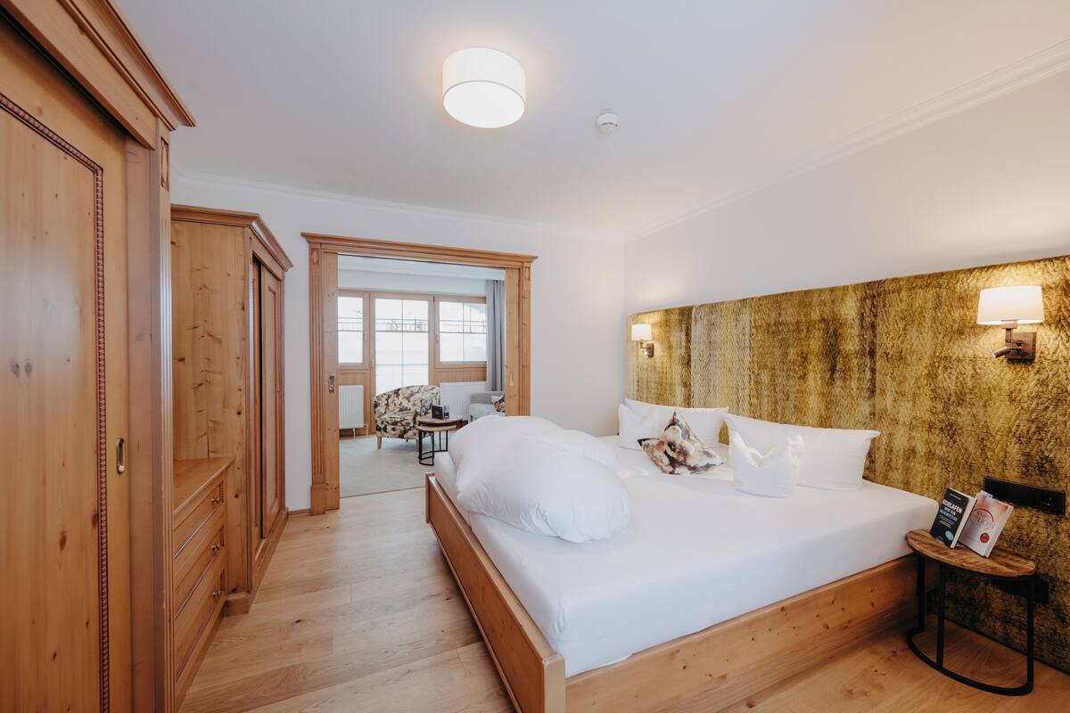 A hotel room with a wooden bed, a headboard with wall-mounted lights, a small table with a lamp, and a view of the outside through a door.