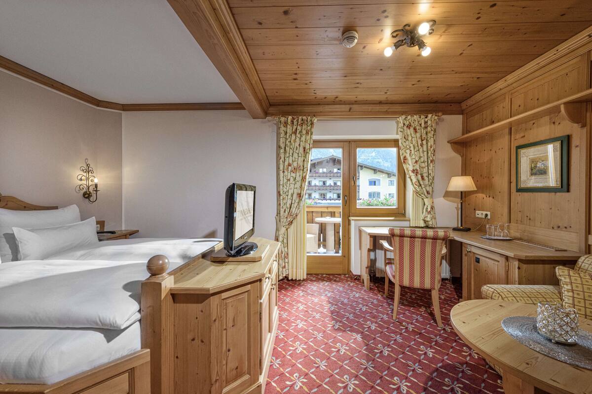 A hotel room with a wooden ceiling, a bed with white linens, a desk with a chair, a small table with a lamp, and a view of buildings through a window with curtains.