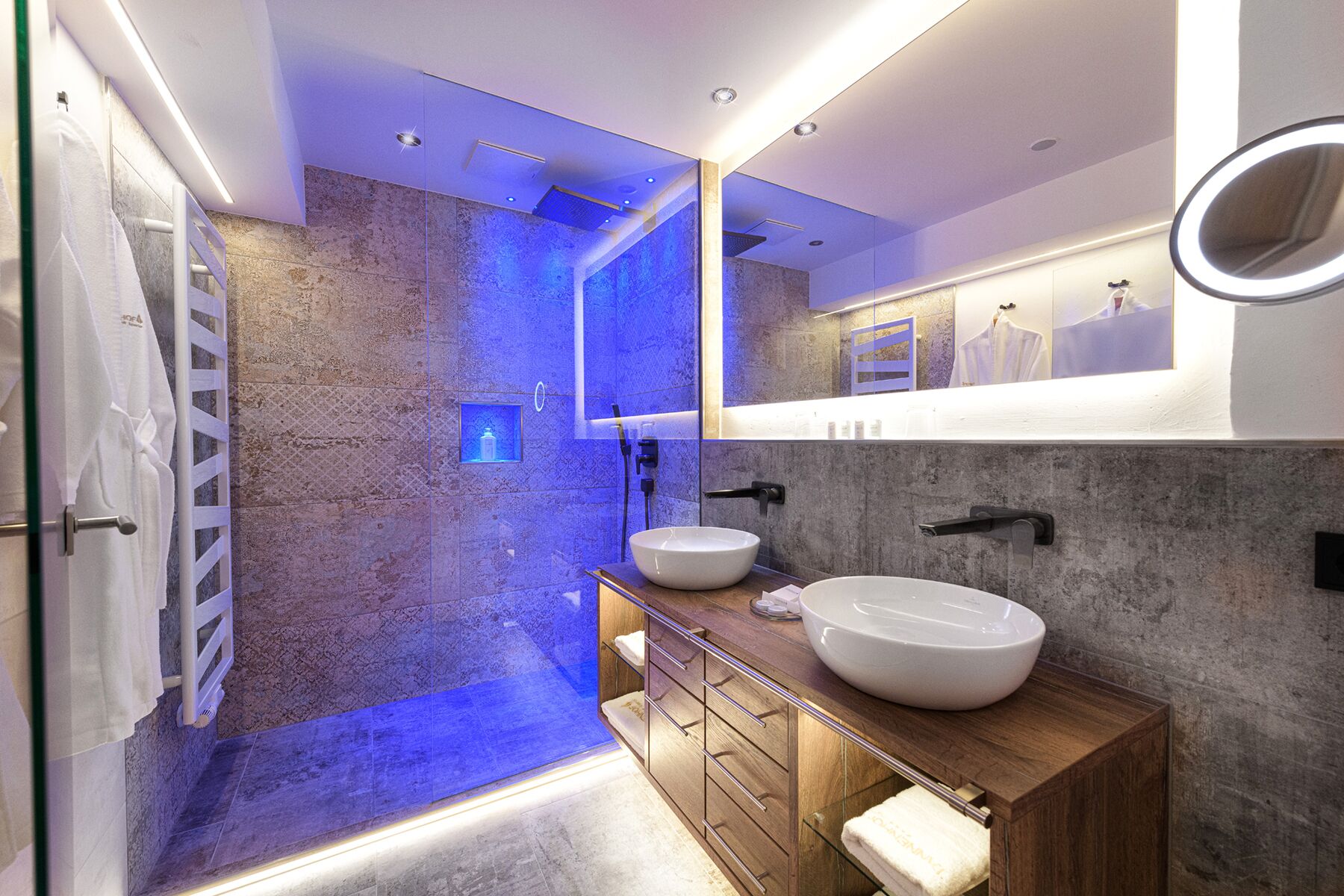 A modern bathroom with a stone wall, a wooden vanity, two sinks, a shower area, and a mirror with a light.