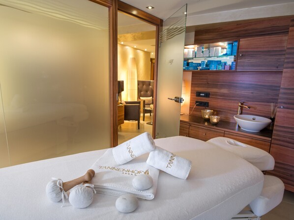 A hotel room with a bed, a bathroom with a sink and mirror, and a massage area with towels and a wooden massage tool.