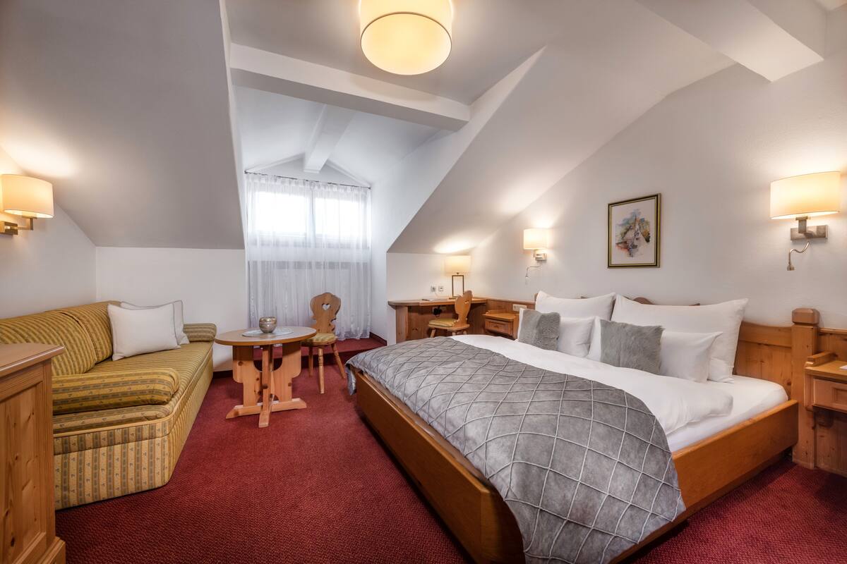A hotel room with a bed, a sofa, a desk, and a chair. There are two lamps on the walls and a painting on the wall.