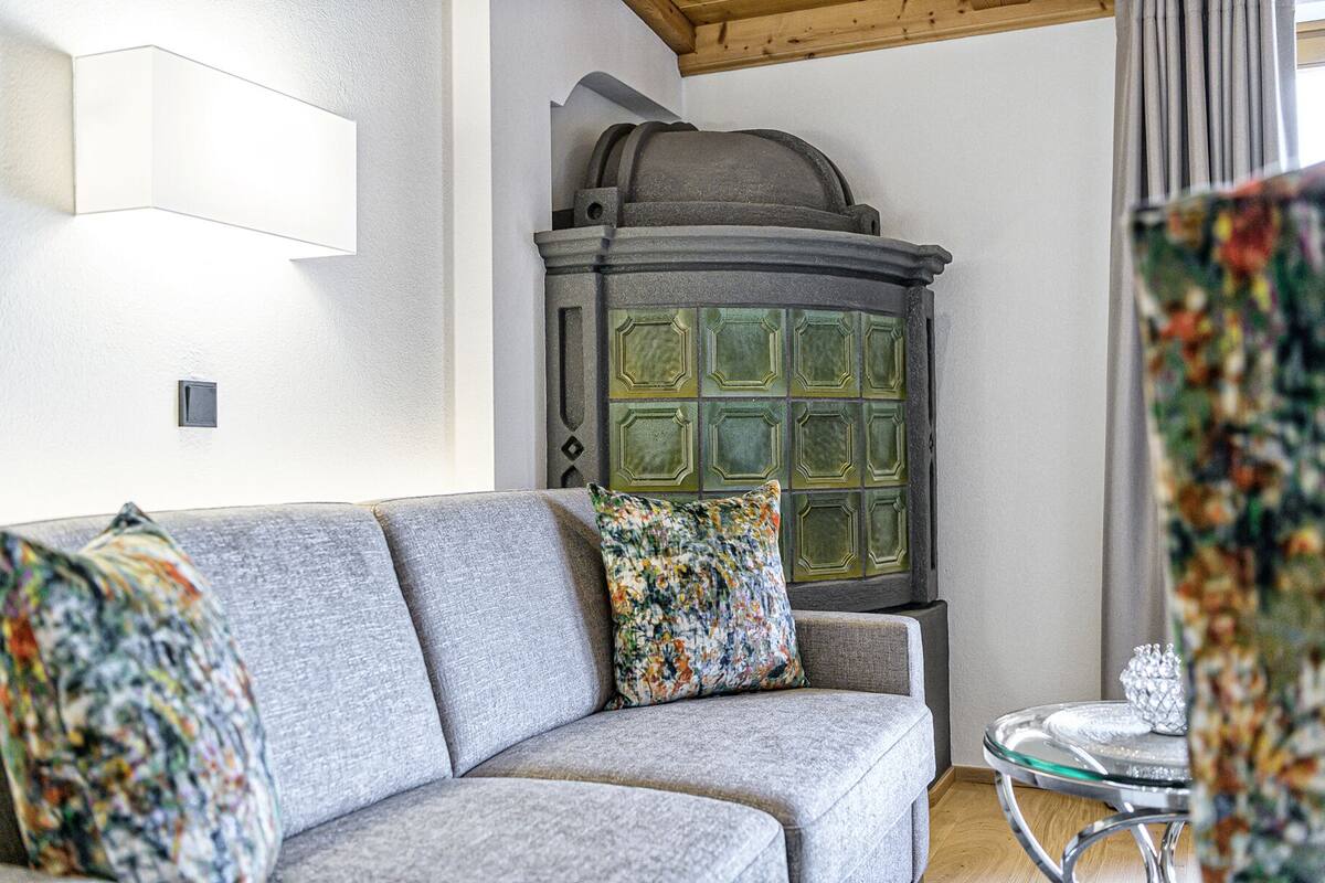 A living room with a grey sofa, a colorful throw pillow, a glass table with a decorative item, and a green tiled stove.