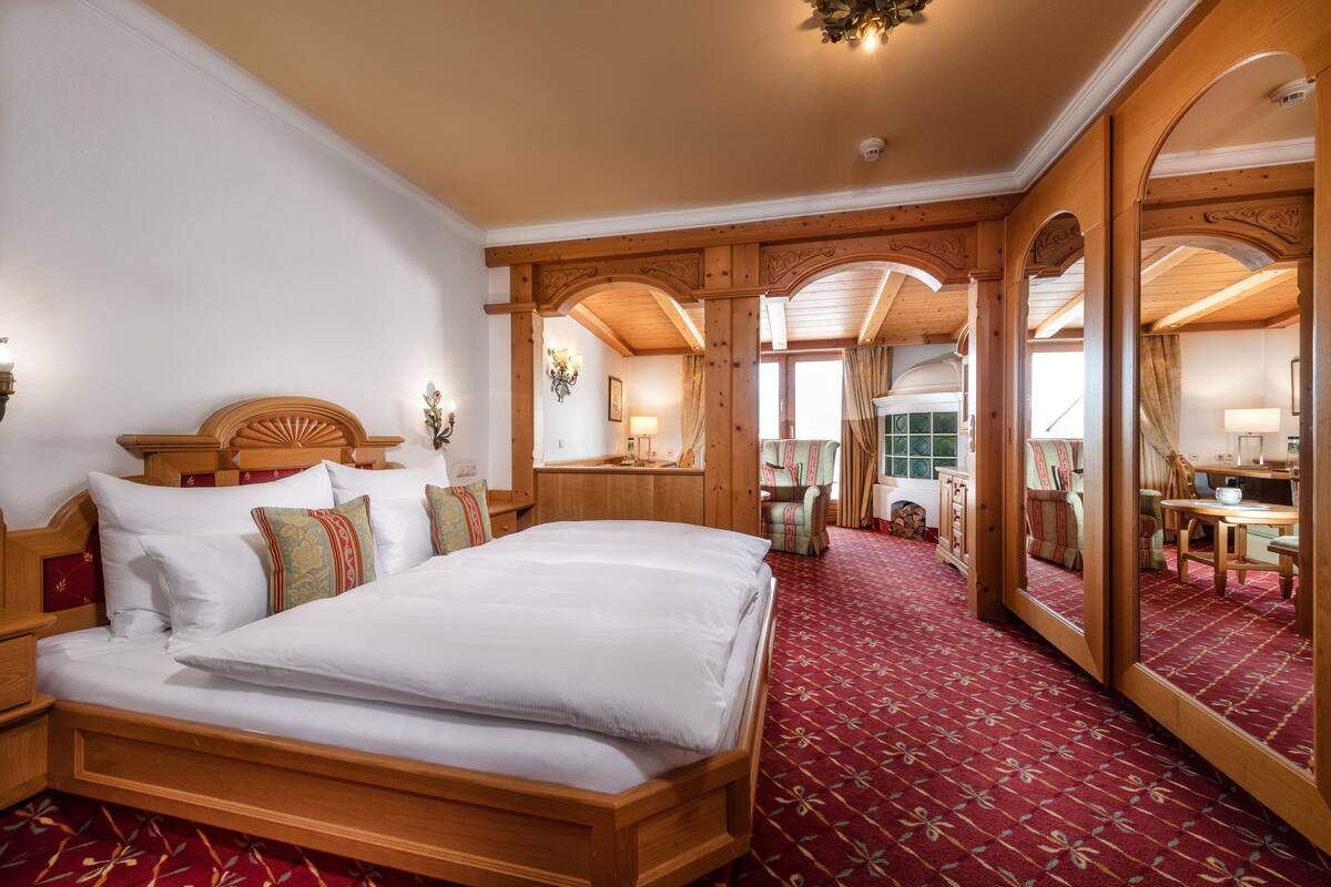 A large wooden bed with white linens, a red patterned carpet, and a large mirror reflecting a dining area.