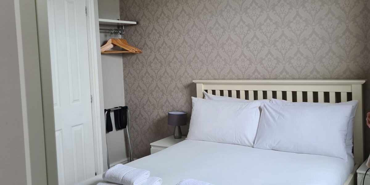 Een net opgemaakt bed met witte lakens, een bedlampje en een houten kapstok aan de muur.