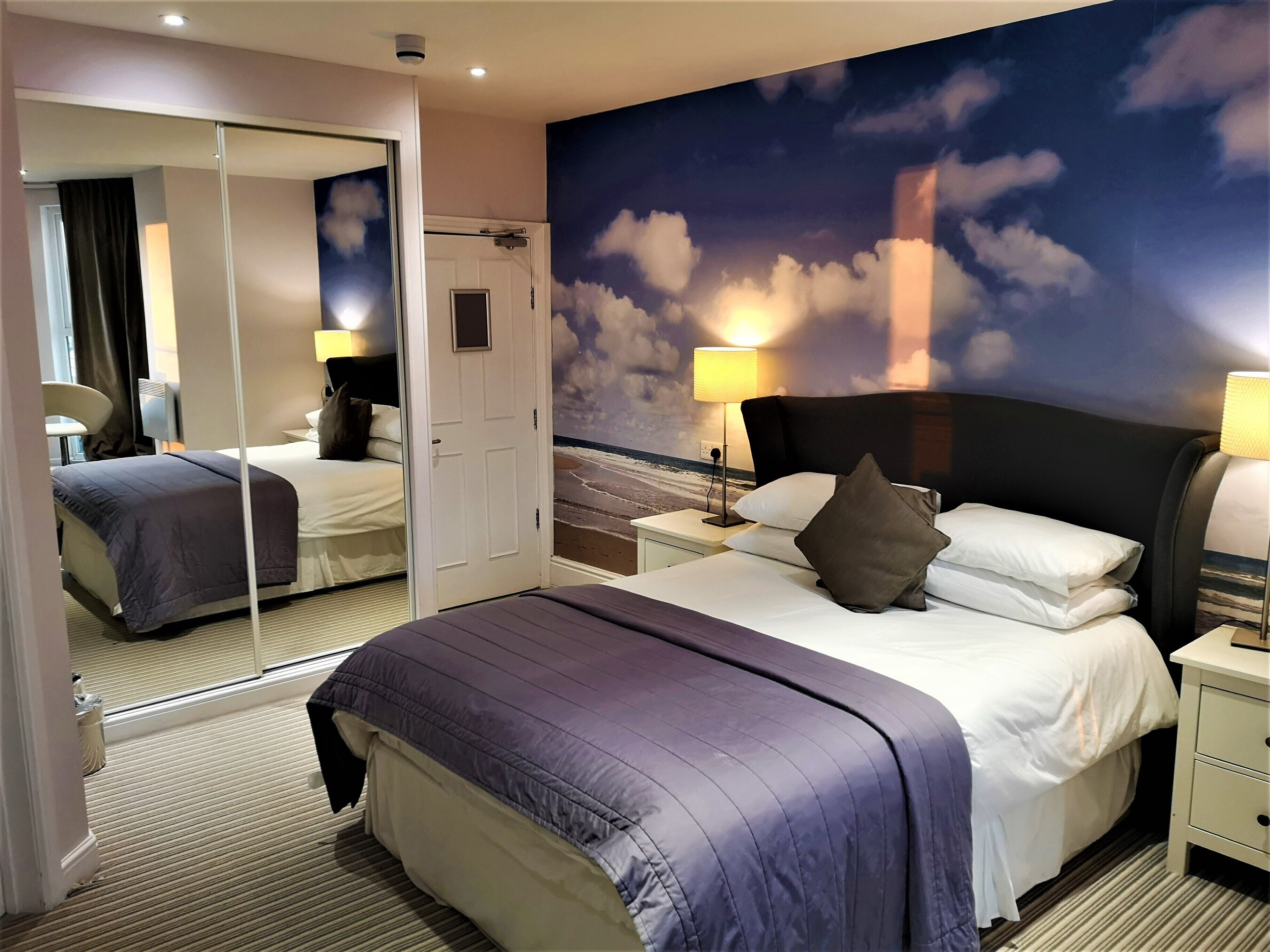 A hotel room with a large bed, a nightstand, a mirror, and a wall mural of clouds.