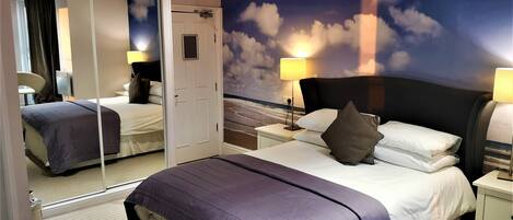 A hotel room with a large bed, a nightstand, a mirror, and a wall mural of clouds.