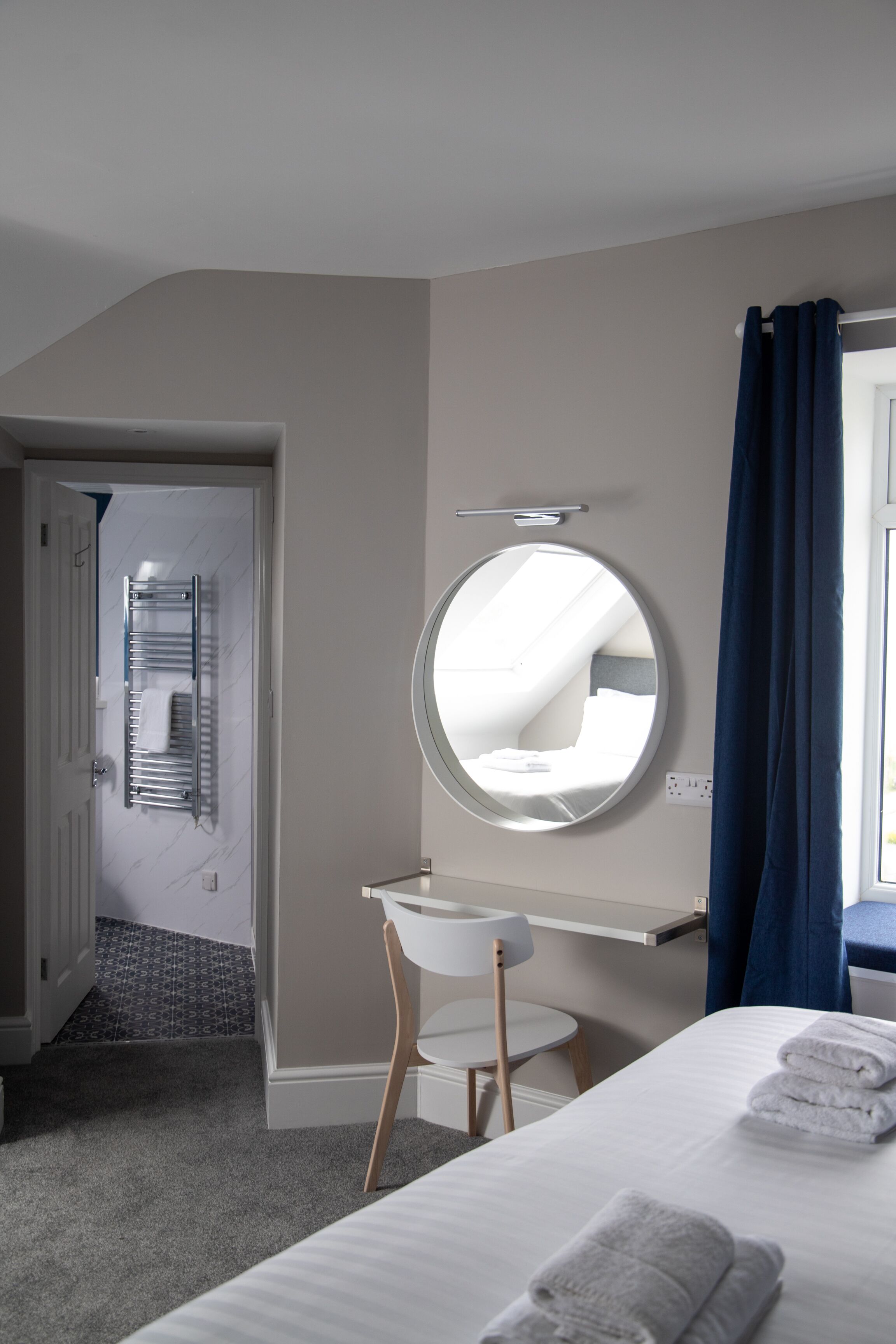 Habitación de hotel con cama, espejo redondo, una pequeña silla blanca, toallero y ventana con cortinas azules.