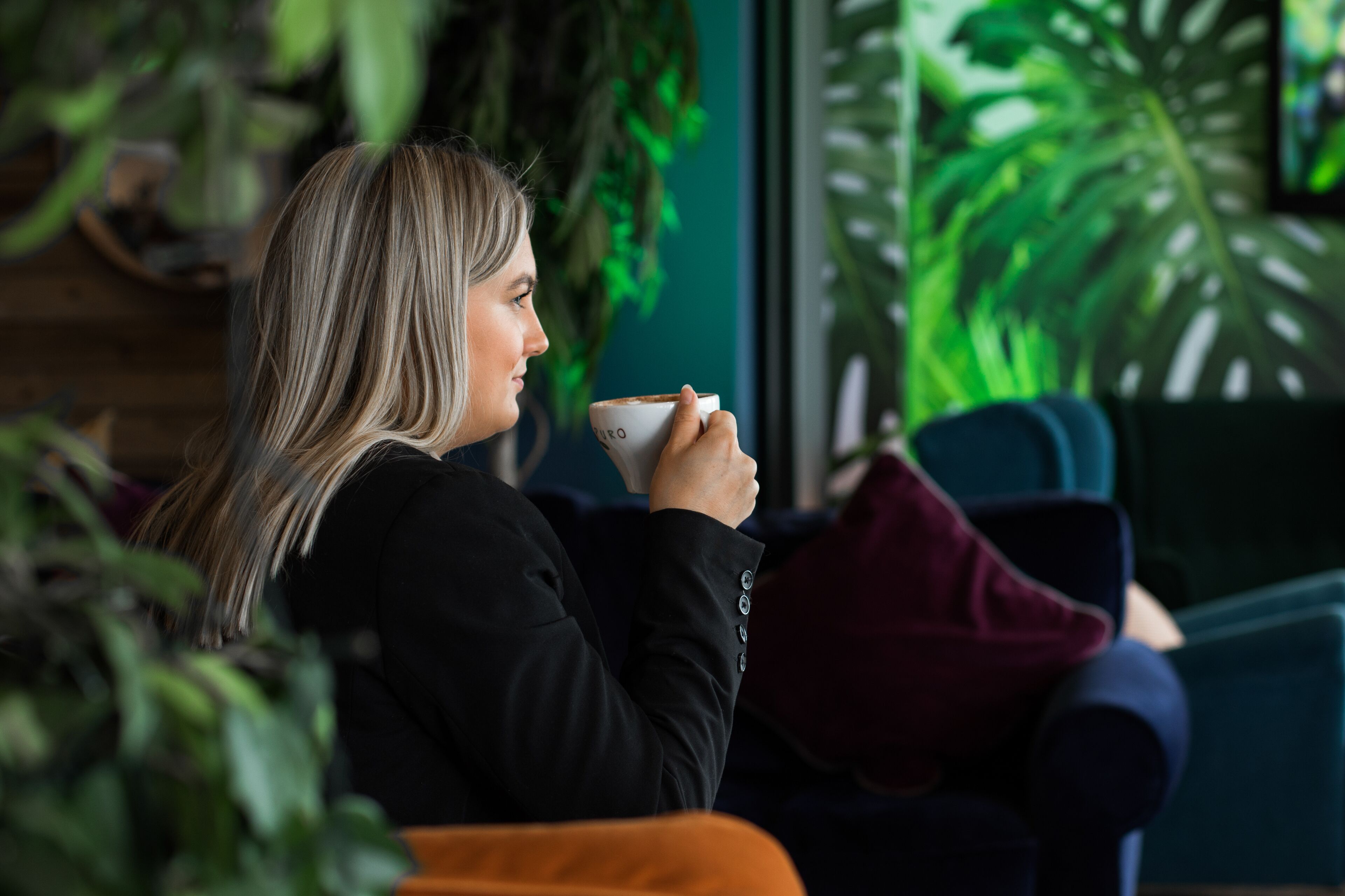 A person sitting on a couch in a room with large windows and indoor plants.