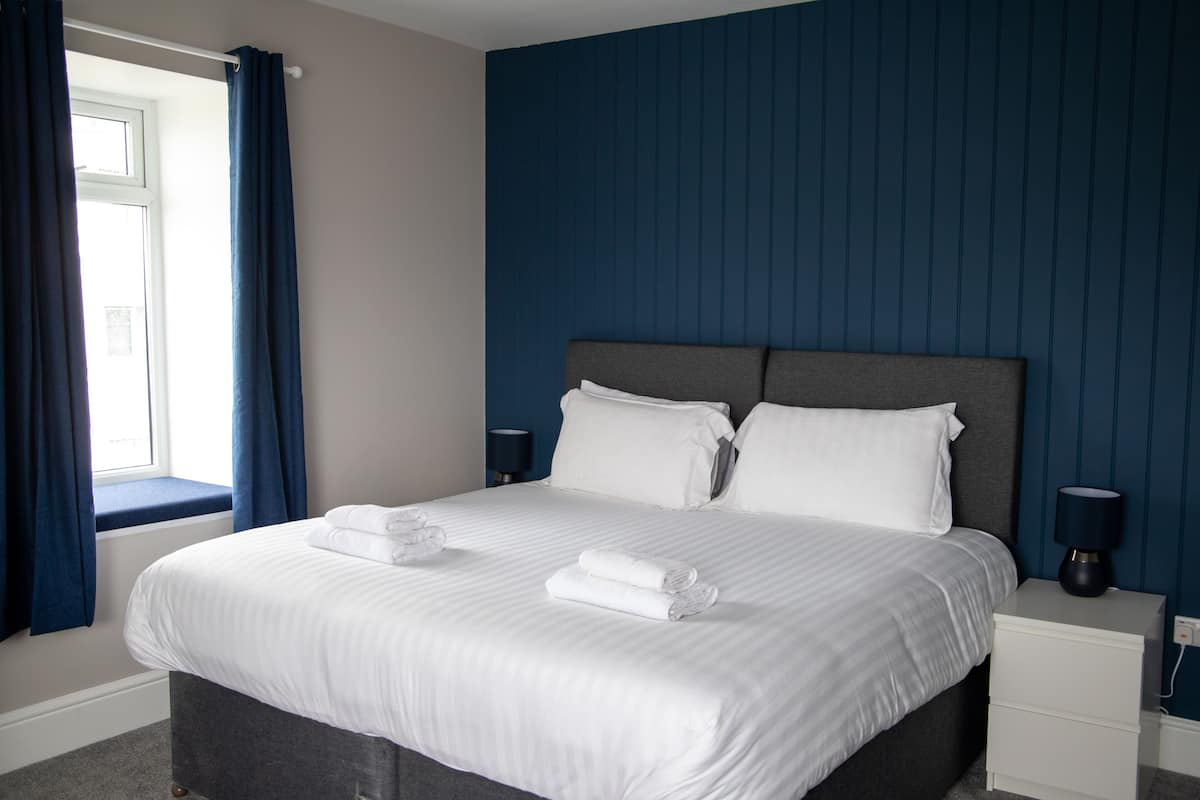 A neatly made bed with white linens and two pillows, a bedside table with a lamp, and a window with blue curtains.