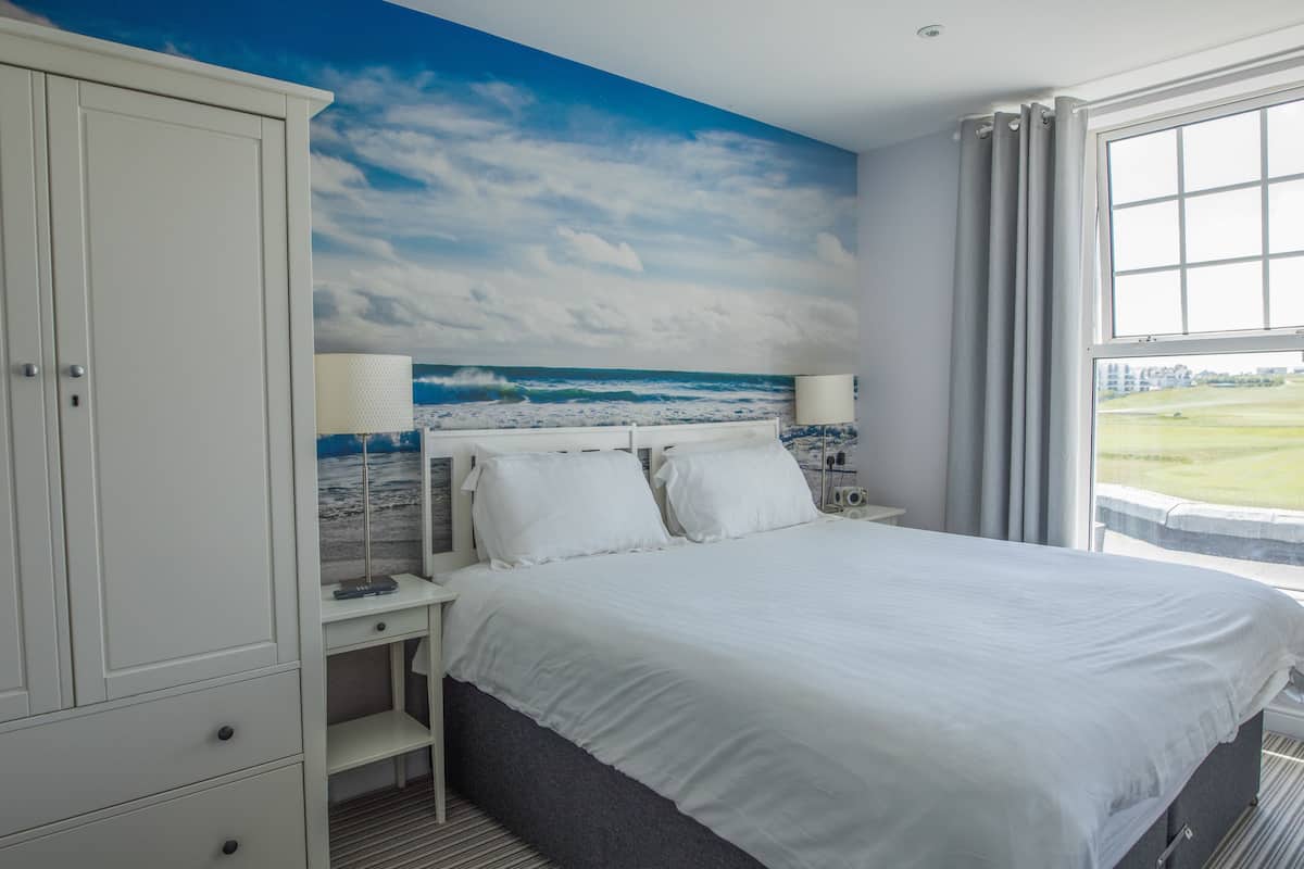 A bedroom with a bed, a nightstand, a wardrobe, a window with a view of a grassy field, and a mural of the ocean on the ceiling.