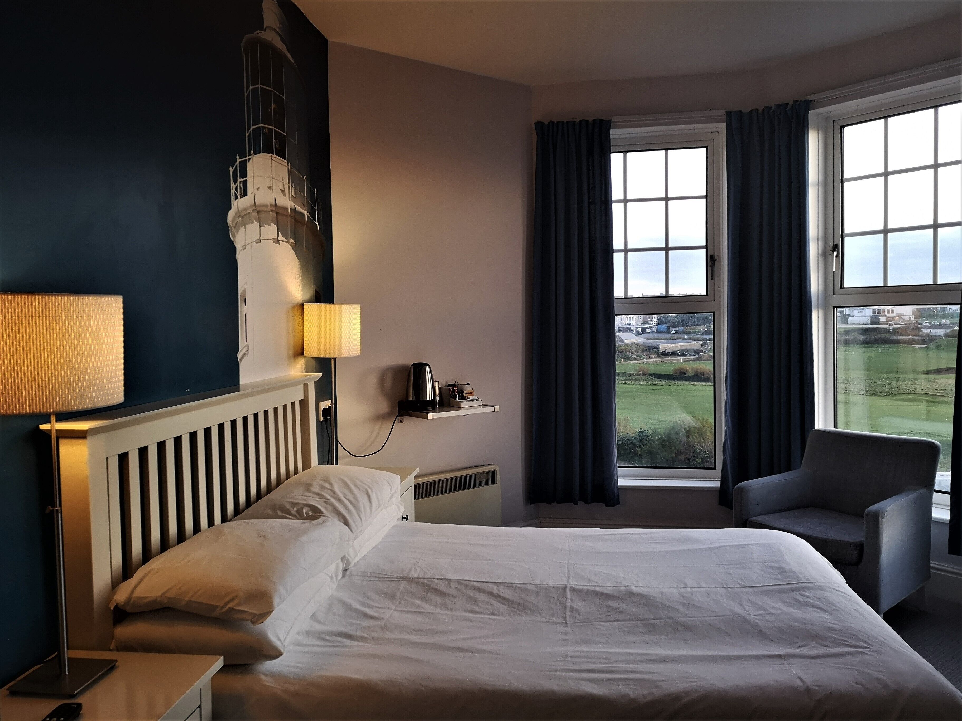 Une chambre d’hôtel avec un lit, une chaise, une lampe, une étagère avec une bouilloire et une vue sur l’extérieur.