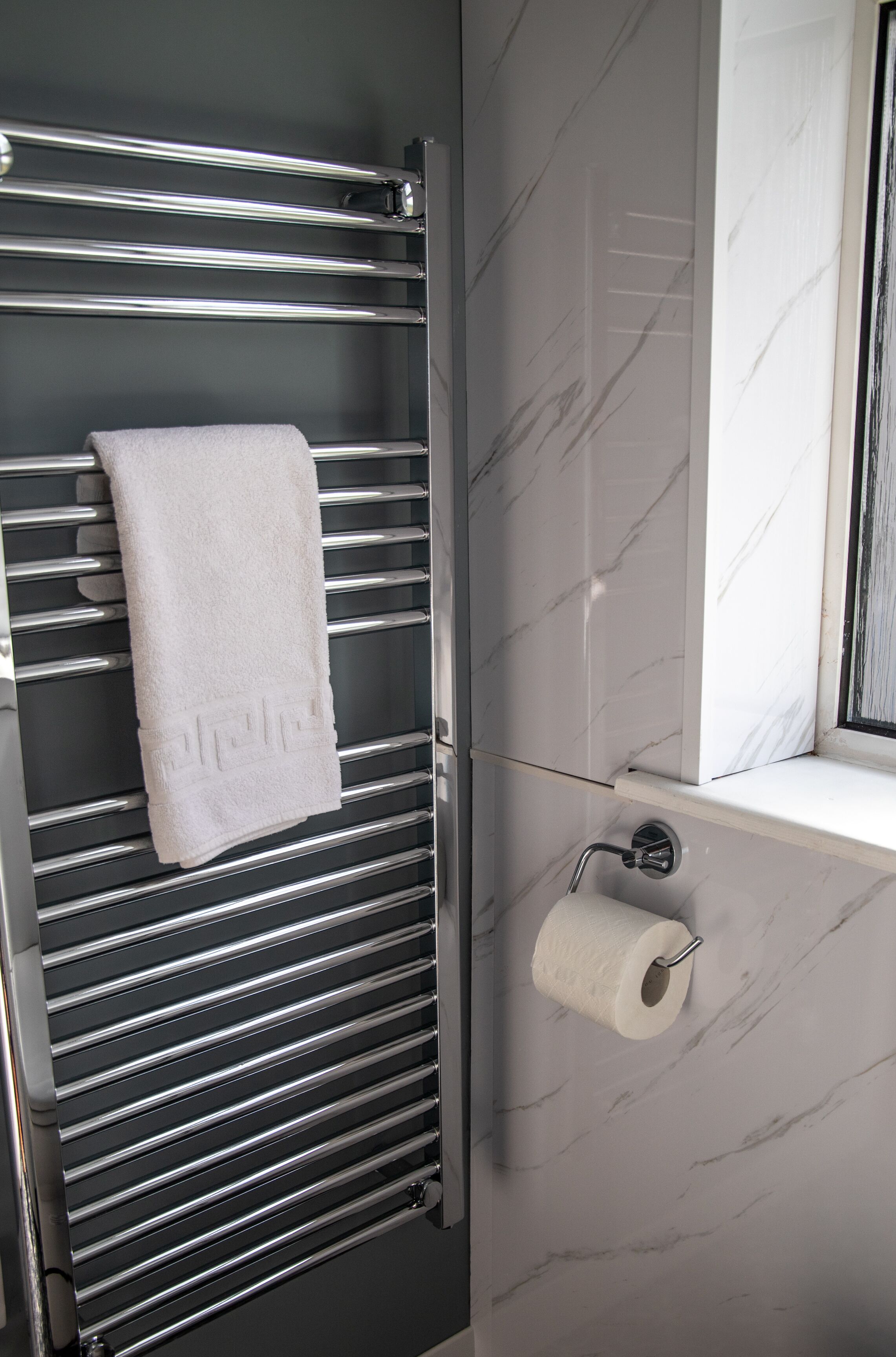 A bathroom with a chrome towel rack, a white towel, marble walls, and a toilet paper holder.