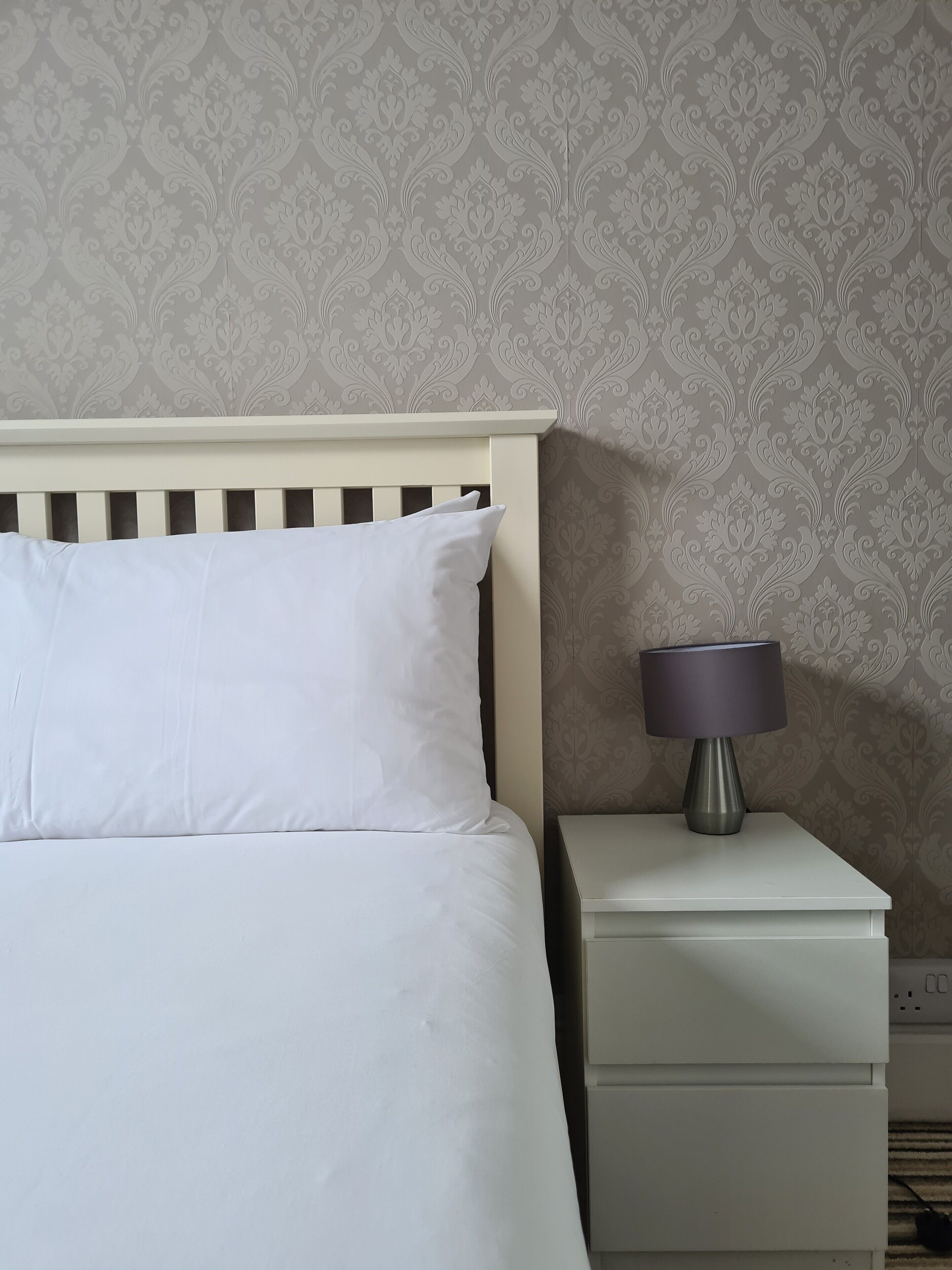 A neatly made bed with white bedding, a white nightstand with a lamp, and a patterned wallpaper.