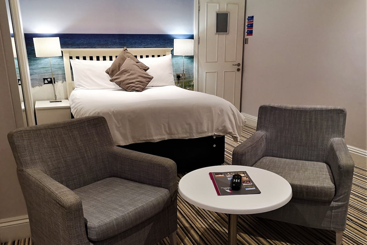 A hotel room with a bed, two armchairs, a small table, and a view of the ocean.