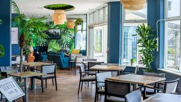 Un café moderne avec des tables et des chaises en bois, de grandes fenêtres et des plantes d’intérieur.