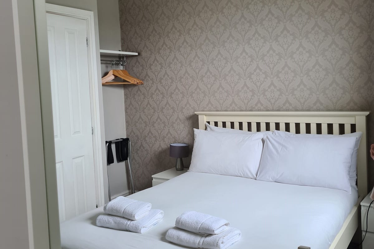 A neatly made bed with white linens and towels, a bedside table with a lamp, and a wardrobe with hanging clothes.