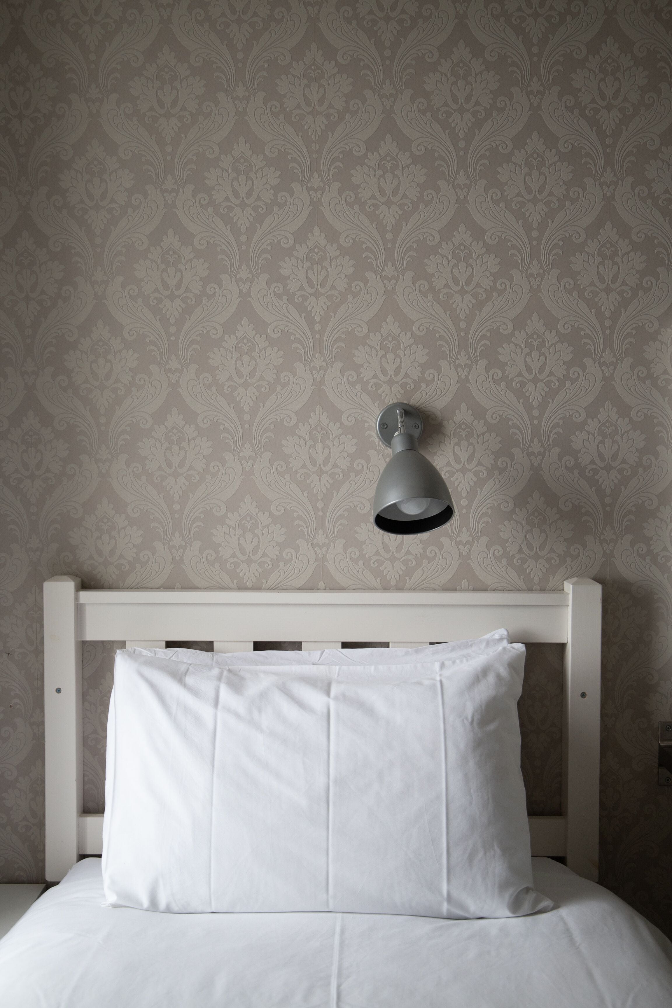 Ein Bett mit weißer Bettwäsche und einem Kopfkissen, ein weißer Kopfteil sowie eine Wand mit floralem Tapetenmuster.