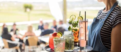 A person holding a tray with various drinks, including a pink cocktail, a red drink, and a green drink with mint leaves.