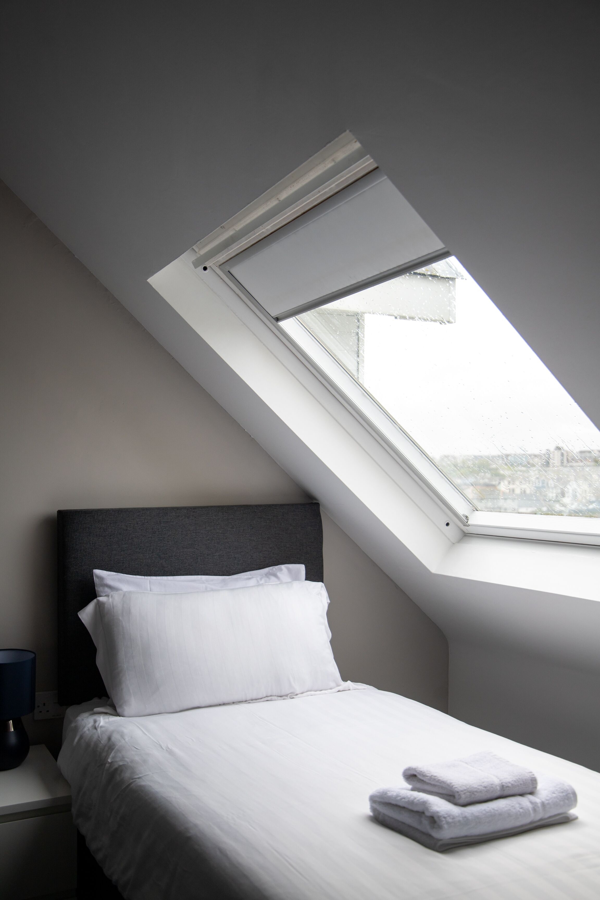 Un dormitorio con tragaluz, cama con ropa blanca y una toalla doblada con cuidado sobre la cama.