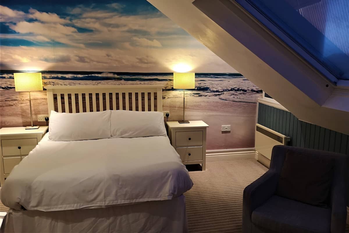 A bedroom with a bed, bedside tables, a lamp, a chair, and a view of the beach through a window.