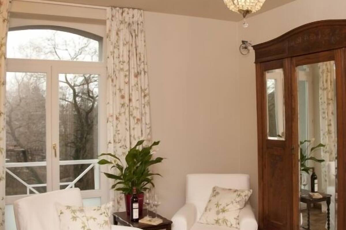 Une pièce avec deux fauteuils blancs, un meuble en bois et une grande fenêtre avec des rideaux.