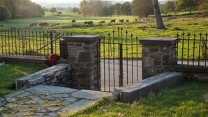 Une porte en pierre, ornée de barreaux métalliques, qui mène à un champ verdoyant où paissent des vaches.