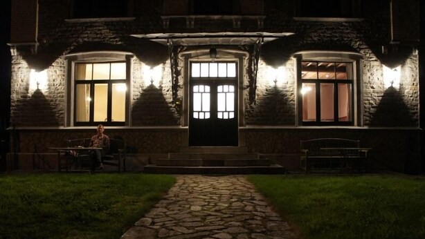 Un bâtiment en pierre avec une entrée centrale, deux fenêtres de chaque côté et un chemin menant à l’entrée.
