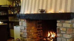 Une cheminée en pierre avec un feu qui crépite, un panier de bois de chauffage et une étagère avec des bouteilles et des verres.